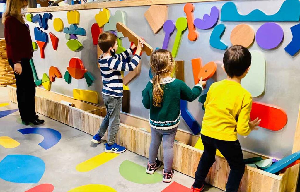 Un gruppo di bambini di spalle che crea opere d'arte sulla parete di gioco utilizzando forme colorate