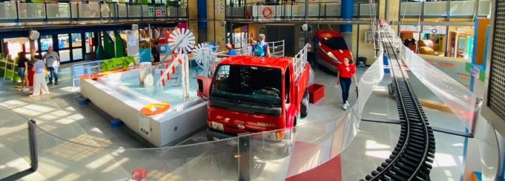 Foto dall'alto della zona centrale di Explora con fontana di giochi d'acqua, treno Frecciarossa 1000 e camion dei pompieri.